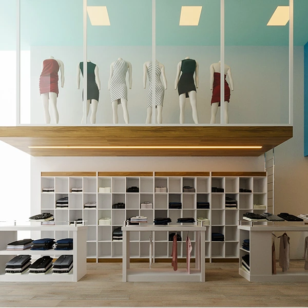 A foto apresenta a decoração de uma loja de roupa pequena e simples, com foco na parede esquerda superior, onde manequins são dispostos de maneira inovadora no teto. Este arranjo peculiar é uma utilização criativa do espaço, chamando a atenção para as peças de vestuário que os manequins exibem. Abaixo, um conjunto de prateleiras e mesas brancas organiza e exibe calçados e acessórios, enquanto a iluminação embutida na estrutura de madeira acima ilumina tanto os manequins quanto os produtos nas prateleiras.